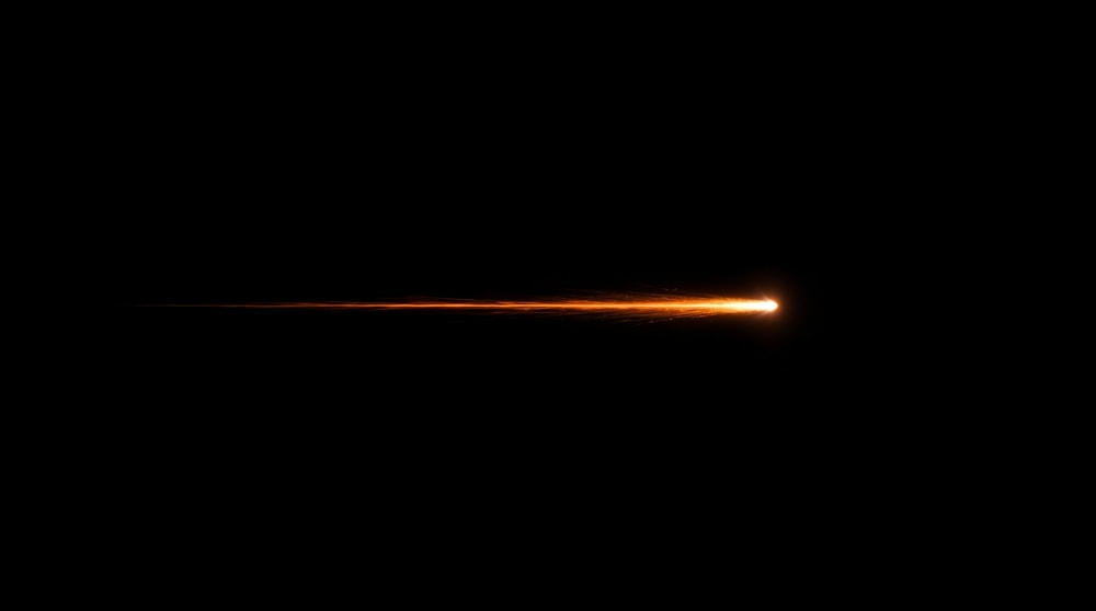 A single, thin shooting star with a bright orange core and a faint, fragmented trail moving horizontally from left to right against a black background.