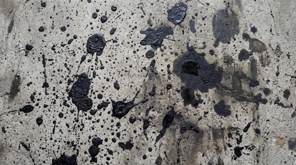 A close-up, top-down view of a raw concrete surface covered in numerous black oil spills and splatters.