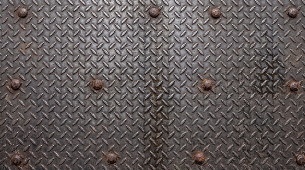 A close-up of a dark industrial diamond plate steel sheet with visible bolt heads.