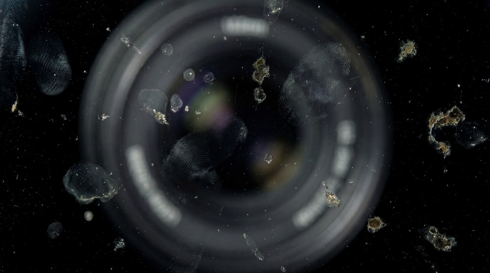 Macro shot of a dirty camera lens with visible fingerprints, smudges, and debris against a black background.
