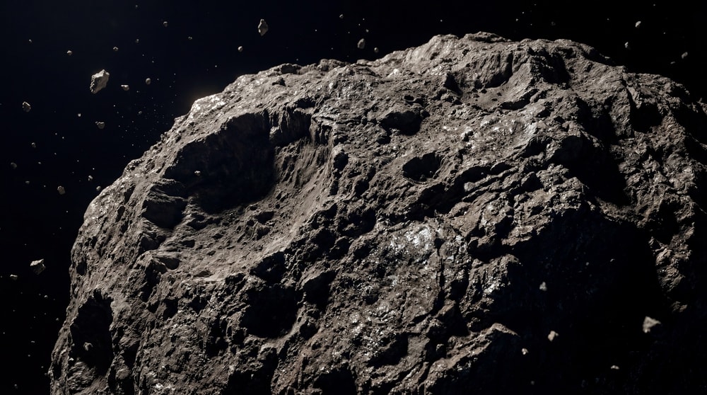 A large, rough, heavily cratered asteroid dominates the foreground, with smaller debris fragments scattered in the dark background.