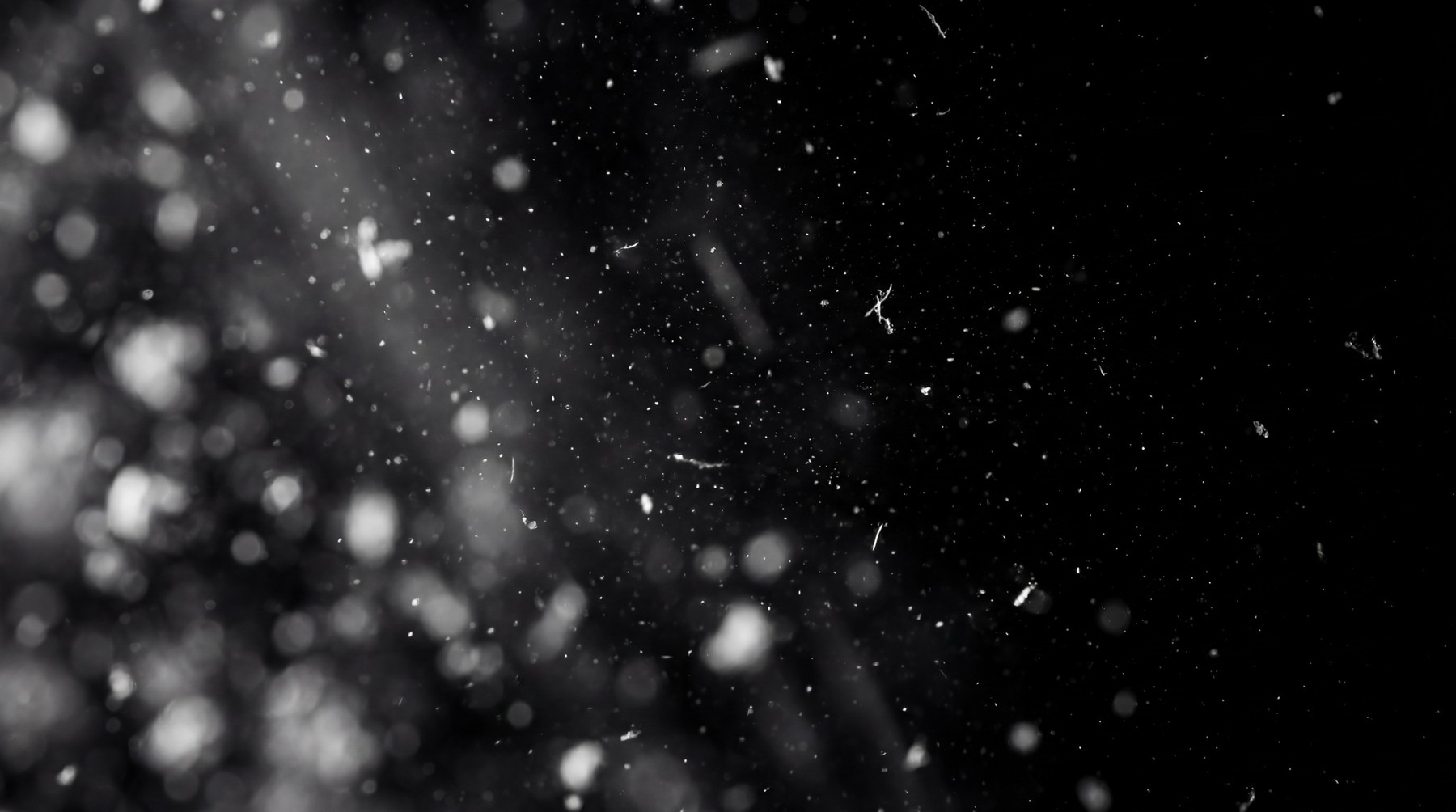 Fine white dust particles scattered against a black background, with some larger bokeh elements on the left and light streaks entering from the top left.