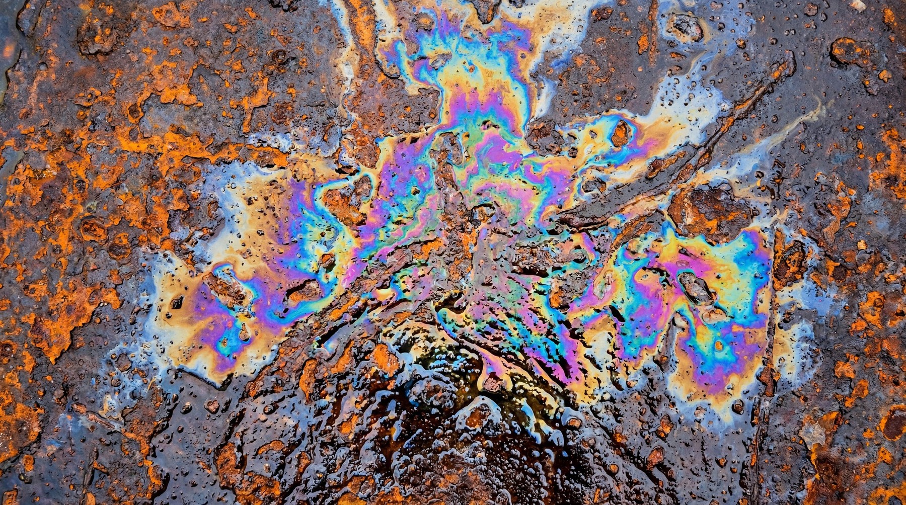 Close-up of a dark, heavily rusted metal surface with a vibrant, iridescent oil slick pattern in the center.