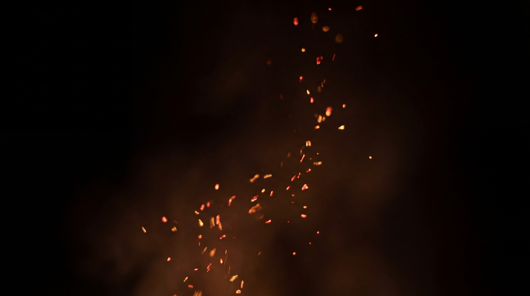 A vertical stream of bright orange and red sparks rising against a dark background with wisps of smoke.