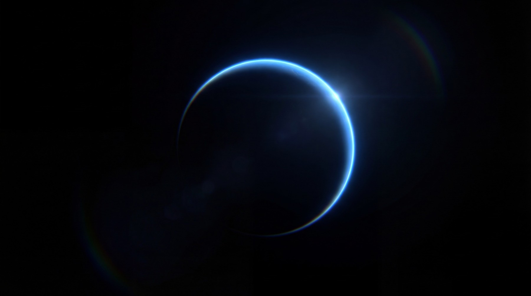 A dark, spherical ice world with a luminous blue halo along its terminator, indicating an atmosphere. Lens flare and subtle rainbow chromatic aberration are visible.