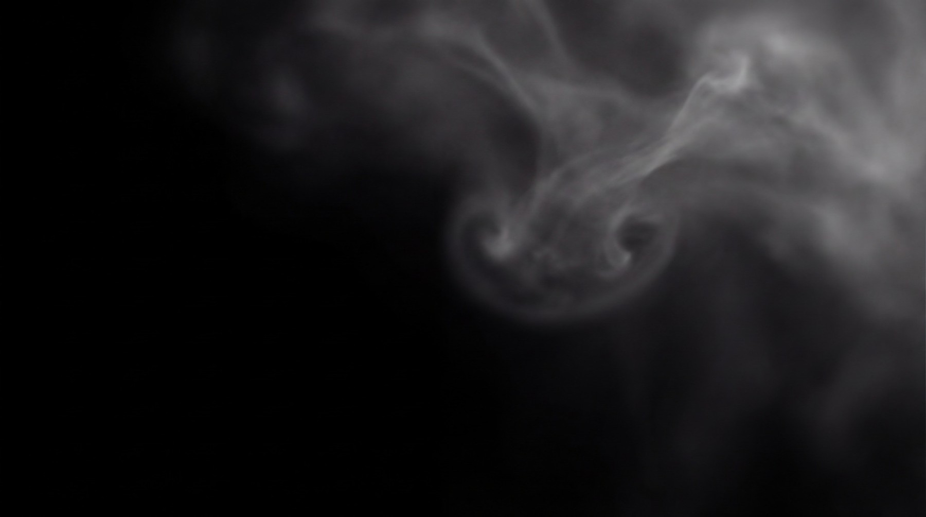 A dark background with a soft, swirling mass of greyish-white vapor forming a distinct circular curl on the right side.