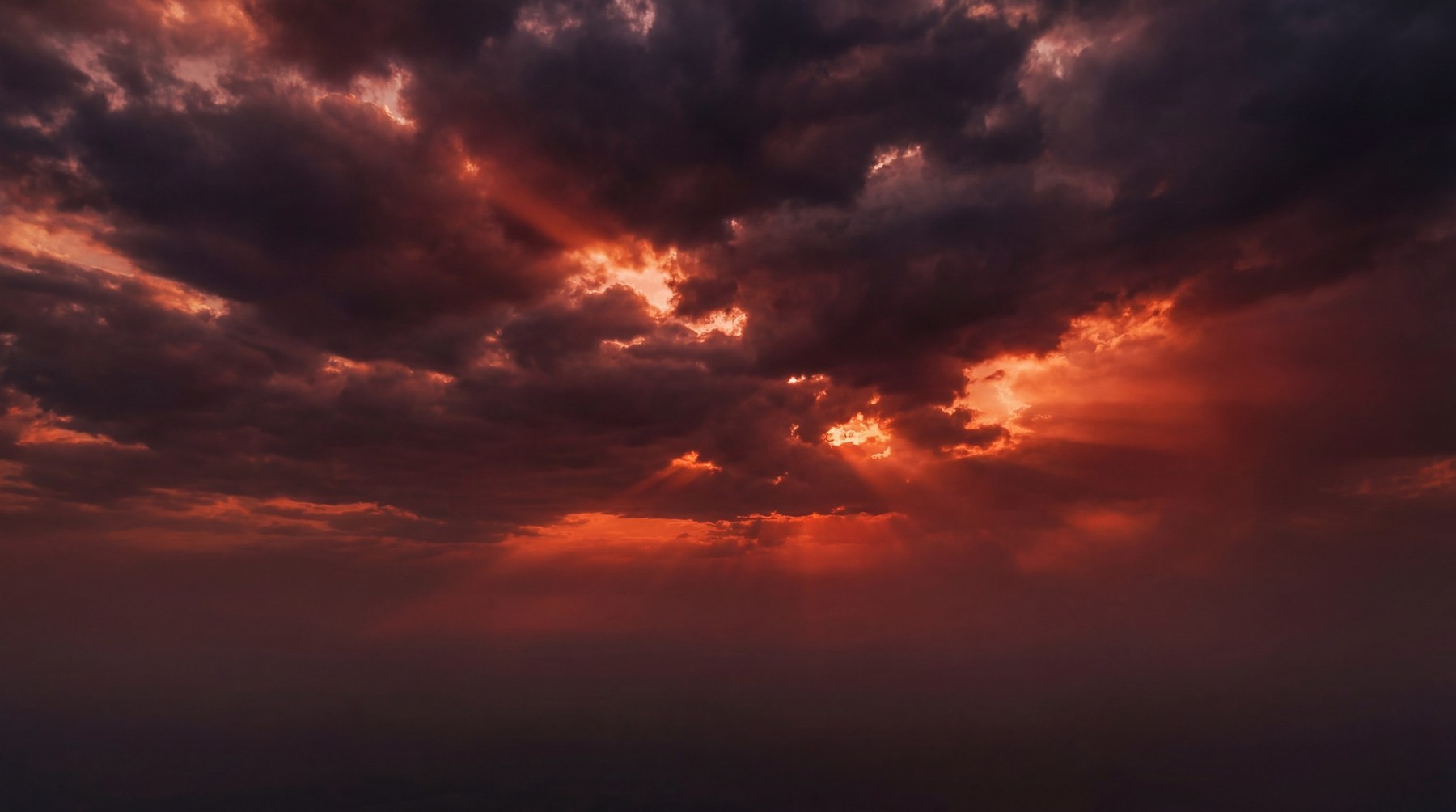 Dramatic sky at sunset with dark, heavy clouds illuminated by intense orange and red light peeking through. Sunbeams radiate downwards.