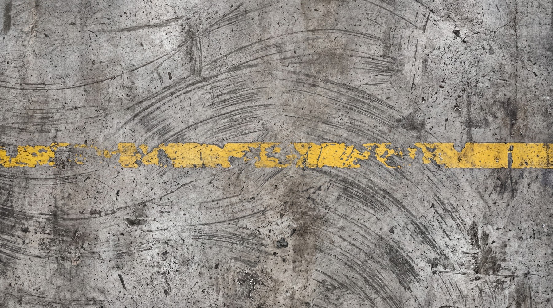 A close-up, top-down view of a textured industrial concrete floor with a broken yellow line painted across it. The concrete surface features numerous scuff marks, tire tracks, and some debris, suggesting heavy use.