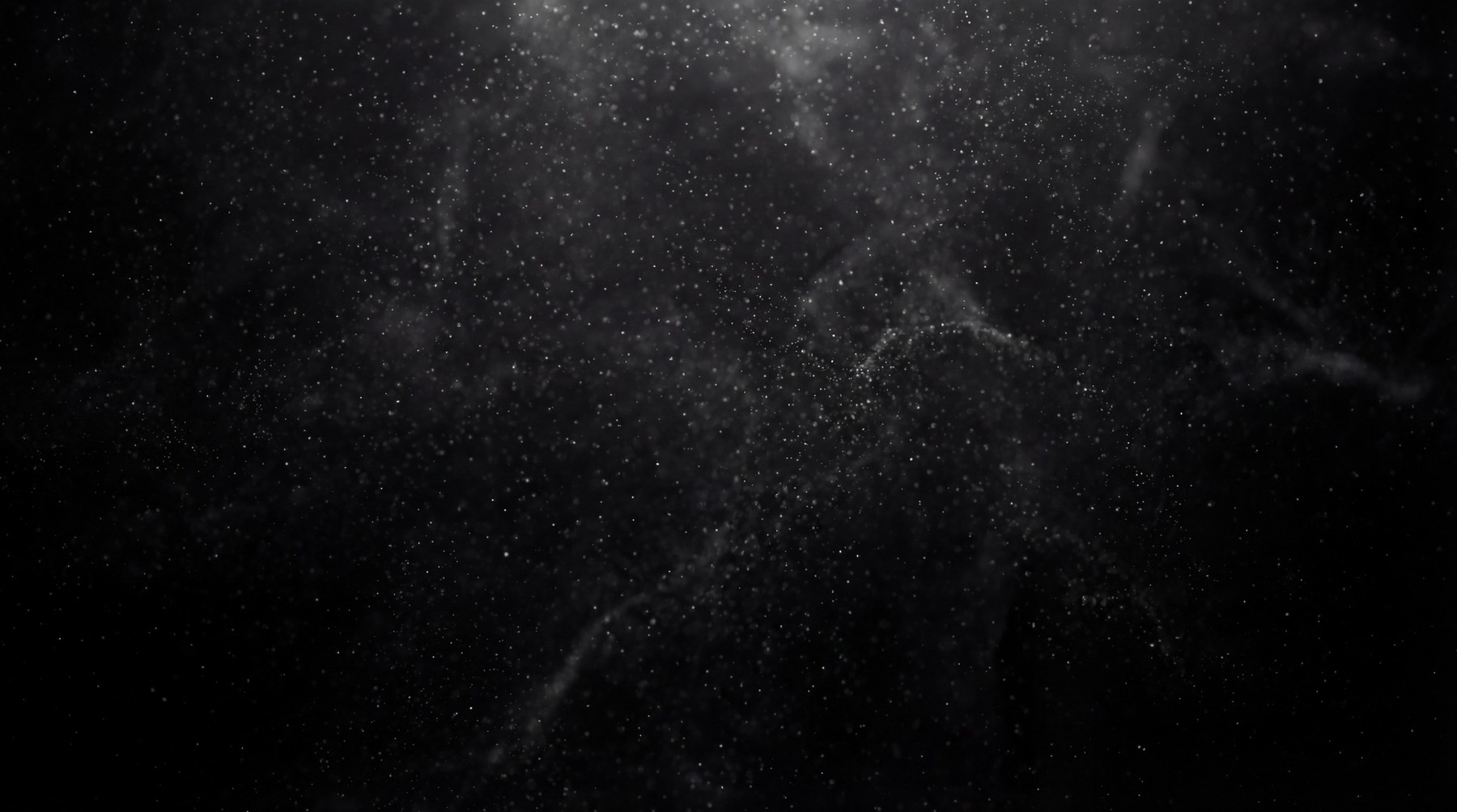 A dark background filled with numerous small, bright white particles resembling cosmic dust or distant stars, with faint wisps of smoke-like texture.