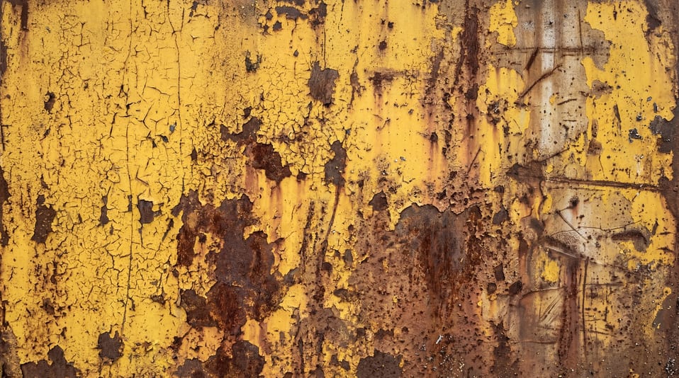 A close-up view of a metal surface with heavily cracked and peeling yellow industrial paint revealing extensive brown rust and oxidized steel patches.