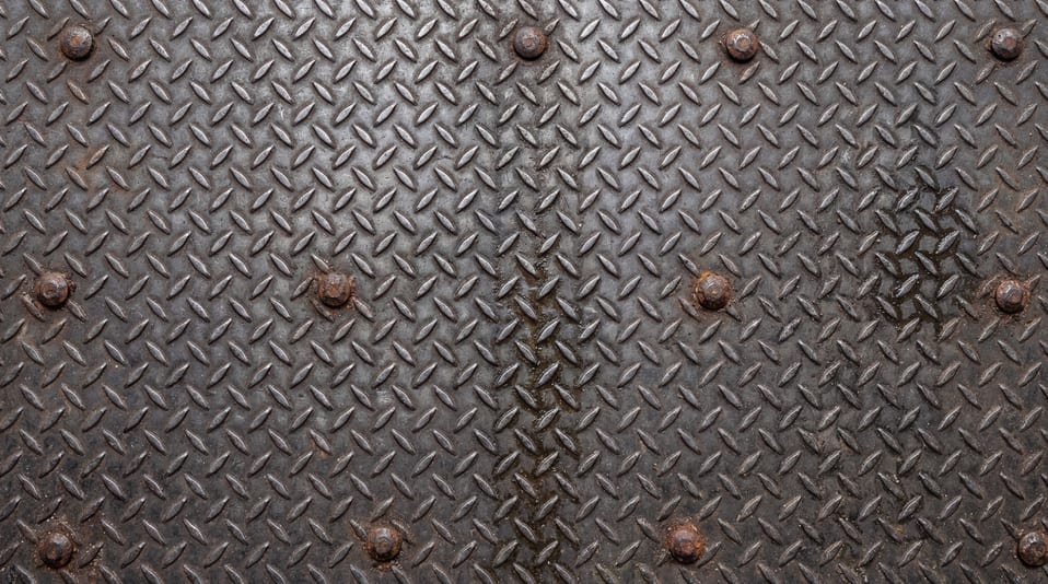 A close-up of a dark industrial diamond plate steel sheet with visible bolt heads.