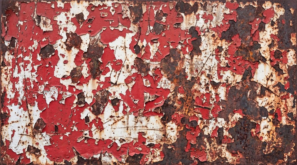 Close-up of weathered steel surface featuring layers of peeling red paint, heavy rust, and deep metallic oxidation.